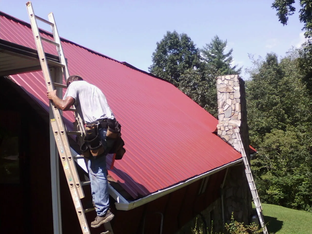 Roofer on a ladder beside a red metal roof and stone chimney — Soffit & Fascia Repair by Roofing Brothers in Donalsonville